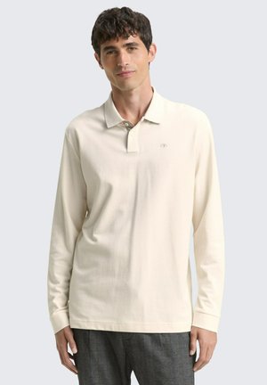 Helle beige Langarm-Polo mit weicher Textur, Kragen und einer Dreiknopf-Leiste. Kleines Logo auf der Brust. Kombiniert mit grauen Hosen.