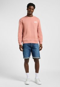 Roze sweatshirt met Lee-logo; denim shorts met opgerolde zomen; witte sneakers; model staat tegen een lichtgrijze achtergrond.