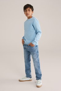 Gaiši zils džemperis, kas izgatavots no kokvilnas, apvienots ar gaiši ziliem džinsiem un baltiem sporta apaviem ar bēša zolēm. Modelis stāv ar rokām kabatās.