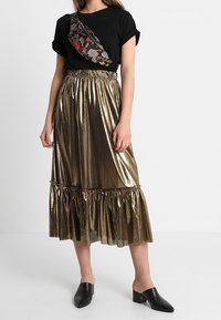 Černé tričko s krátkým rukávem a květinovými detaily, v kombinaci se lesklou zlatou sukní střední délky s volánkovým lemem. Černé nazouvací boty.