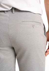 Pantaloni grigi chiari realizzati in tessuto testurizzato, con una tasca posteriore dotata di un dettaglio con bottone. Design semplice e aderente, che mette in evidenza la zona vita.