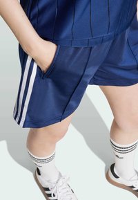 Marina atletiska shorts med vita sidostripes och fickor, tillverkade av slätt tyg, kombinerat med vita strumpor med svarta ränder och sneakers.