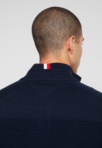 Homme aux cheveux courts vu de dos, portant un pull en maille bleu marine avec un petit détail en rayures rouge, blanc et bleu sur le col.