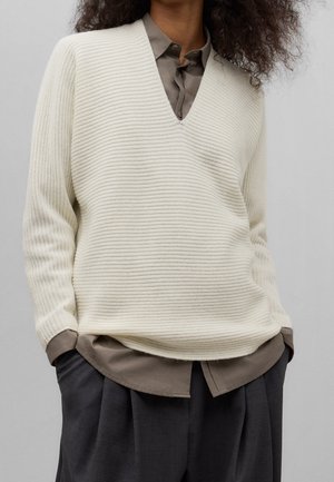 Personne portant un pull à col en V côtelé crème sur une chemise taupe boutonnée, un pantalon ample gris foncé et les mains dans les poches.