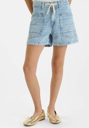 Femme portant un short en denim bleu clair avec de grandes poches avant et un cordon blanc, associé à des ballerines dorées.