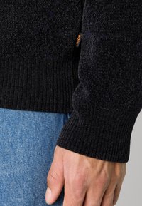 Gros plan sur une main portant un pull à manches côtelées noir avec une petite étiquette orange "BOSS" au-dessus d'un jean en denim bleu.