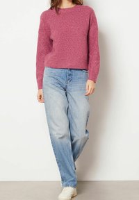 Pull tricoté rose avec un motif en relief en forme de losanges, col rond et manches longues, porté avec un jean bleu clair ample et des baskets blanches.