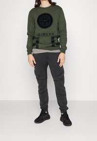 Persoon in een olijfgroen sweatshirt met zwart bijenembleem en de tekst "GLORIOUS", zwarte cargobroek en zwarte sneakers, staande op een grijze vloer.