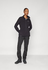 Svart fleecejakke med høy krage og glidelås foran, kombinert med svarte cargo-bukser med lommer og en avslappet passform. Svart fottøy fullfører utseendet.