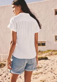 Camicia bianca a colletto con maniche corte, abbinata a pantaloni corti in denim blu chiaro. La camicia presenta un dettaglio arricciato sul retro e una vestibilità casual.