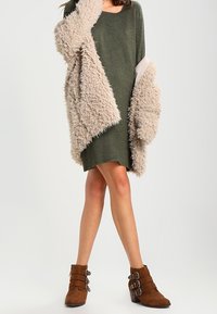 Olivgrön klänning med lös passform, lager på lager med en beige fuskpäls stor sjal, och bruna ankelboots med spänne.