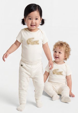 Deux tout-petits en tenues crème assorties avec des logos crocodile dorés, l'un debout et l'autre assis, tous deux souriants sur un fond blanc.