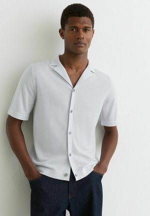 Mężczyzna stoi oparty o białą ścianę, ubrany w białą koszulę na krótki rękaw z guzikami oraz ciemnoniebieskie dżinsy, z rękami w kieszeniach.