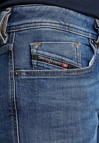 Jeans de mezclilla azul oscuro con un bolsillo trasero que lleva una etiqueta negra y roja de "Diesel Industry", acentuada con costuras doradas y un detalle de remache.