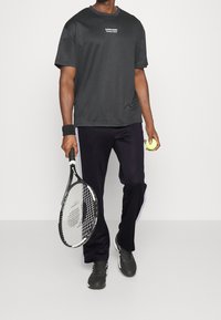 Černé funkční tričko s logem, spárované s námořnickými sportovními kalhotami s bílými bočními pruhy. Drží tenisovou raketu a míček.