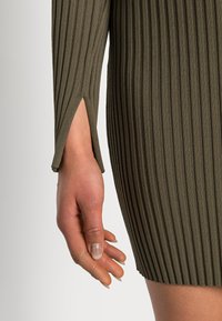 Olivgrön, ribbad klänning med lång ärm och sidoklor. Tyget verkar mjukt med en texturerad finish som framhäver konturer.