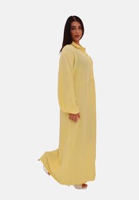 Lungo vestito giallo realizzato in tessuto strutturato, con chiusura frontale a bottoni e maniche lunghe. Cade fino alle caviglie con una vestibilità ampia.