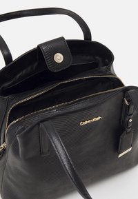 Sort håndtaske med en tekstureret overflade, der har dobbelte håndtag, en kliklås og en guldfarvet lynlås. Indvendigt er der flere rum.