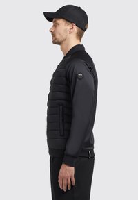 Giubbotto puffo nero con maniche in tessuto liscio, colletto a coste e tasche con zip. Presenta una patch con il logo sulla manica sinistra.