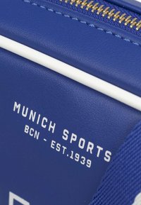 Borsa sintetica blu con accenti bianchi, dotata di zip dorata e testo in rilievo: "MUNICH SPORTS BCN - EST. 1939." Finitura testurizzata.
