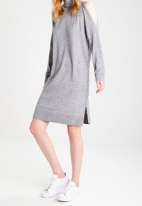 Robe grise en maille avec manches longues et épaules dénudées. Comprend des fentes latérales et un ourlet côtelé, portée avec des baskets blanches.