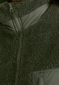 Chaqueta de forro polar en verde oscuro con textura suave, cierre frontal con cremallera, cuello alto y un detalle de tejido liso en contraste en el bolsillo.