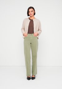 Ανοιχτό μπεζ ζακέτα, καφέ εφαρμοστό τοπ, πράσινο τζιν με ψηλή μέση και ίσιο πόδι, και μαύρες γόβες με μυτερή μύτη, στέκεται μπροστά σε λευκό φόντο.