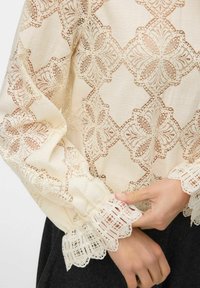 Ecru bluzka z misternym koronkowym wzorem w kwiaty, falbaniastymi mankietami oraz miękkim, teksturowanym materiałem.