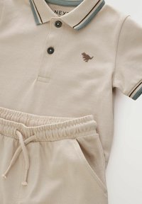 Polo beige avec une finition texturée, présentant une broderie de dinosaure marron foncé, des boutons noirs, et un col à rayures bleu et marron.