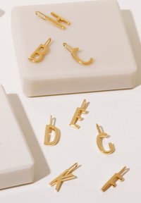 Guldbrevcharms i olika stilar: B, C, D, E, F, G, H och K. Satsen är placerad på en neutralfärgad, slät låda.