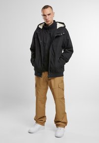 Chaqueta negra aislante con capucha, con forro de felpa blanca, combinada con pantalones cargo beige y zapatillas blancas.