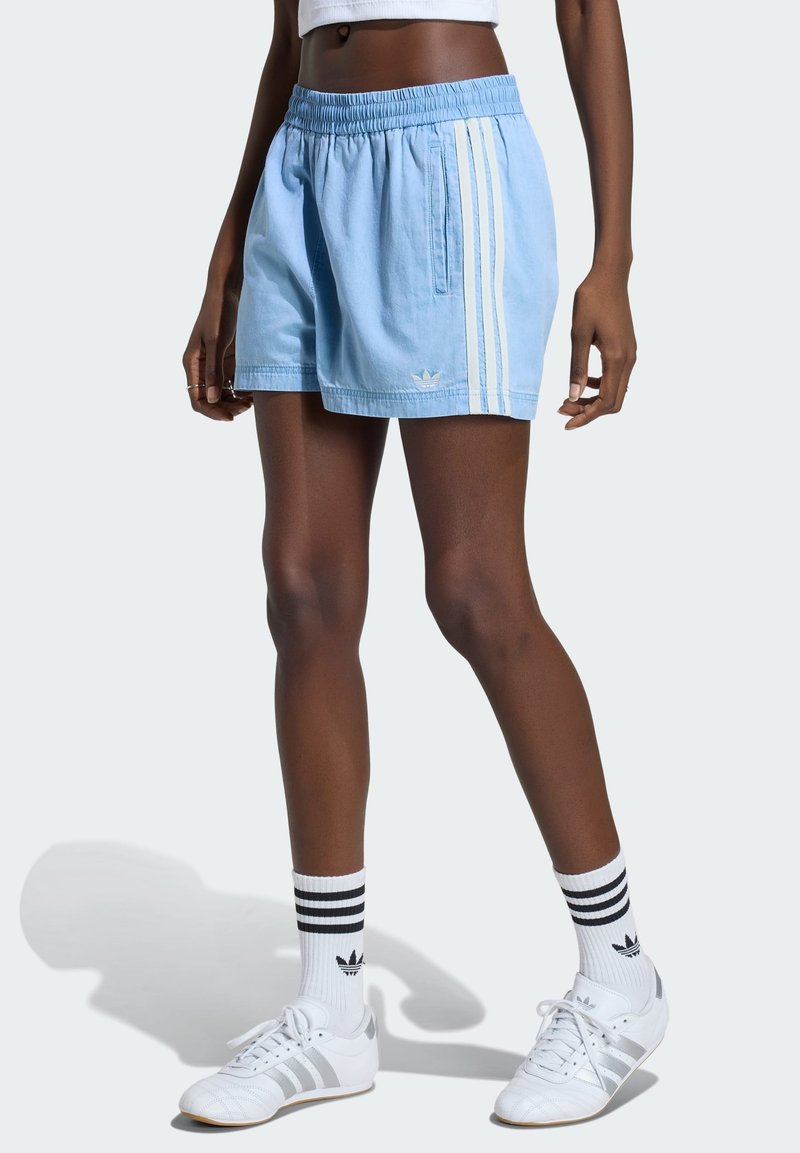 Γυναίκα που φοράει ανοιχτό μπλε σορτς Adidas με λευκές ρίγες, λευκές κάλτσες Adidas με μαύρες ρίγες και λευκά αθλητικά παπούτσια Adidas.