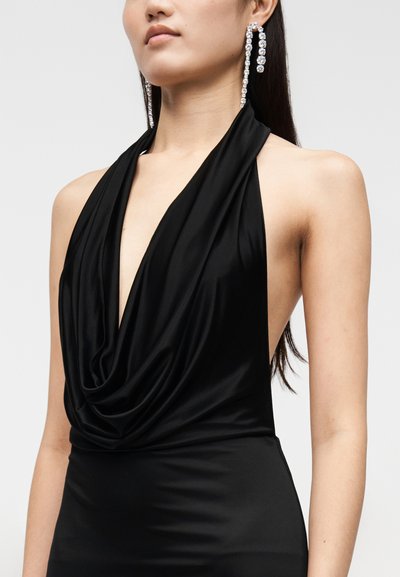 Nő ujjatlan fekete pántos ruhában, fodros nyakkivágással és hosszú kristály csepp fülbevalókkal, sima háttér előtt.