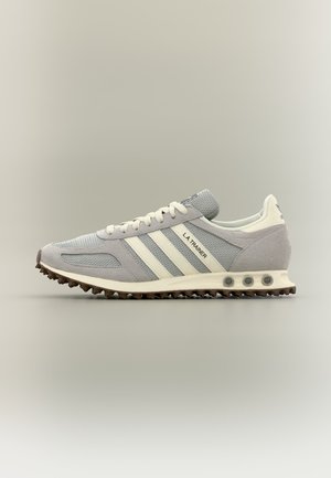 Hellgrauer Adidas L.A. Trainer Sneaker mit weißen Streifen, strukturierter Mesh-Oberfläche und brauner genoppter Sohle auf neutralem Hintergrund.