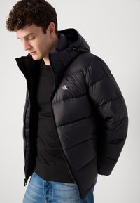 Czarna pikowana kurtka puchowa z kapturem, zapięciem na zamek błyskawiczny i małym haftowanym logo na piersi. Noszona na czarnej koszuli.
