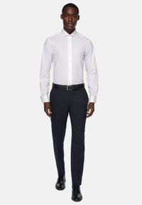 Bela srajca z dolgimi rokavi in strukturiranim ovratnikom, v kombinaciji s temno modrimi hlačami in črnimi poslovnimi čevlji, s klasičnim krojem in gladkim materialom.