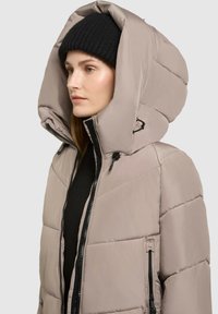 Puffjacka i ljusbeige med stor huva, dragkedja fram, texturerad yta och sidofickor. Svart stickad mössa som bärs på huvudet.