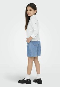 Camisa de manga larga blanca con patrones de corazones rojos, combinada con pantalones cortos de mezclilla azul y zapatos negros, exhibida sobre un fondo claro.