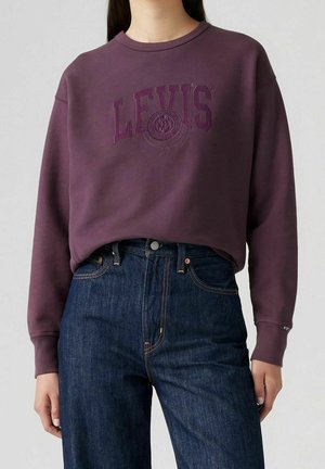 Kobieta w fioletowej bluzie Levi's z tłoczonym logo i ciemnoniebieskich dżinsach z wysokim stanem, stojąca na jednolitym tle.
