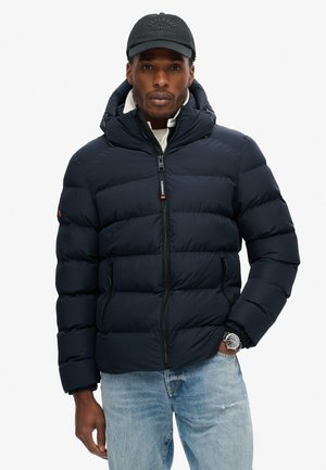 Chaqueta de plumas azul marino con capucha, con secciones acolchadas horizontales, cremallera frontal y dos bolsillos laterales. Lleva puesta una camisa ligera.