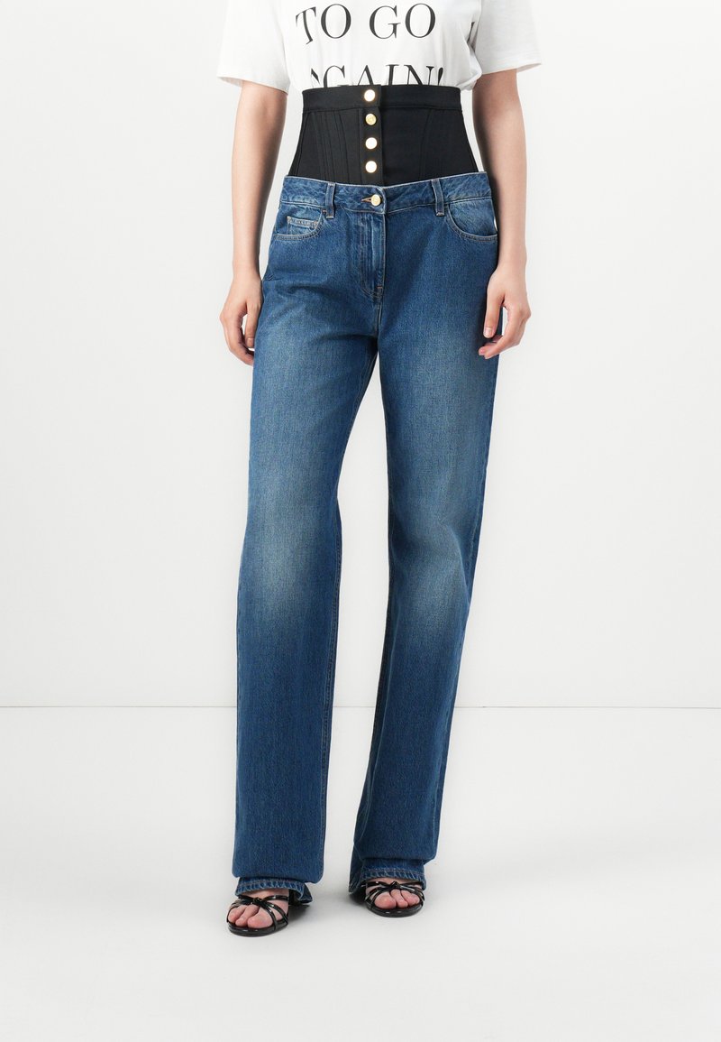 Zwarte hoge tailleband met drie gouden knopen gecombineerd met lichtblauwe denim jeans met rechte pijpen. De stof heeft een gladde textuur.