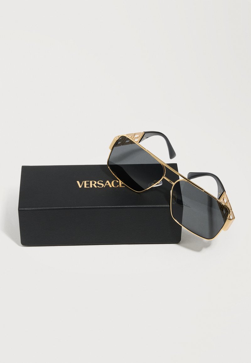 Gold-framed dark lens sunglasses with black temples resting on a black Versace branded box against white background.