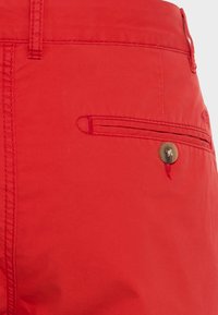 Shorts en coton rouge avec détails de couture, une poche latérale à fermeture par bouton, et une texture lisse.