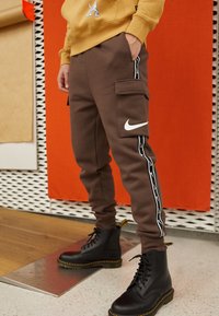 Bruna Nike-joggingbyxor med sidostråk och cargofickor. Kombineras med svarta snörstövlar med guldsömmar.