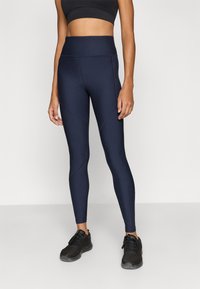 Μπλε αθλητικό κολάν με ψηλή μέση, κατασκευασμένο από υλικό με υφή και ελαστικότητα, με διαγώνιες ραφές, συνδυασμένο με μαύρα αθλητικά παπούτσια.