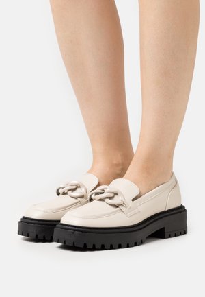 Mocassins beiges avec un accent chaîne, tige en cuir lisse et une épaisse semelle noire à plateforme dotée d'un motif de bande de roulement rugueux.