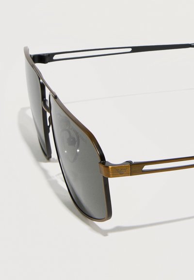 Ορθογώνια γυαλιά ηλίου με σκουρόχρωμους φακούς και λεπτά μπρούτζινα μεταλλικά σκελετά, με κομμένα πλαϊνά μπράτσα και διακριτικό λογότυπο της μάρκας στη μεντεσέ.