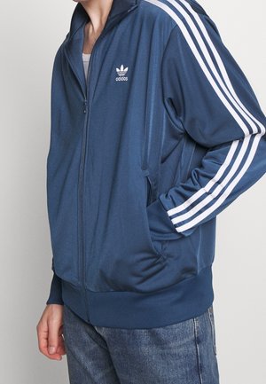 Osoba nosząca niebieską kurtkę adidas na zamek z białymi paskami na rękawach i logo na piersi, w połączeniu z niebieskimi dżinsami.