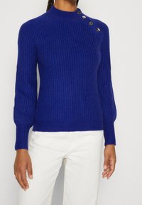 Pull tricoté d'un bleu vif avec un col montant, une texture côtelée et des boutons décoratifs sur l'épaule. Coupe ajustée avec manches longues.