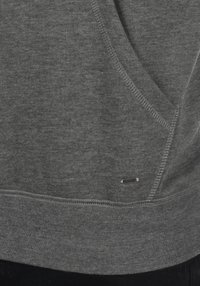 Sweat-shirt gris en tissu doux, avec une poche kangourou, des poignets et un ourlet côtelés, et une étiquette en métal argenté sur le côté.